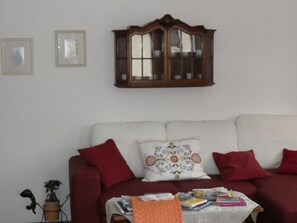 A living room with a white sofa, red and patterned cushions, a wooden cabinet, and a small table with books and a plant. - Chasa Balcun, (Scuol) by Interhome (Scuol)