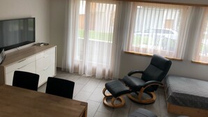 Une pièce comprenant un téléviseur posé sur un meuble en bois, une table à manger avec des chaises, un fauteuil à bascule noir, un lit et de grandes fenêtres ornées de rideaux légers.