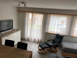 A room with a TV on a wooden cabinet, a dining table with chairs, a black rocking chair, a bed, and large windows with sheer curtains. - Chasa Mundin ****, (Scuol). by Interhome (Scuol)