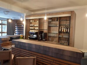 A bar area with a wooden counter, shelves with glasses and bottles, and a coffee machine. - Vvf Villages Prapoutel (Les Adrets)