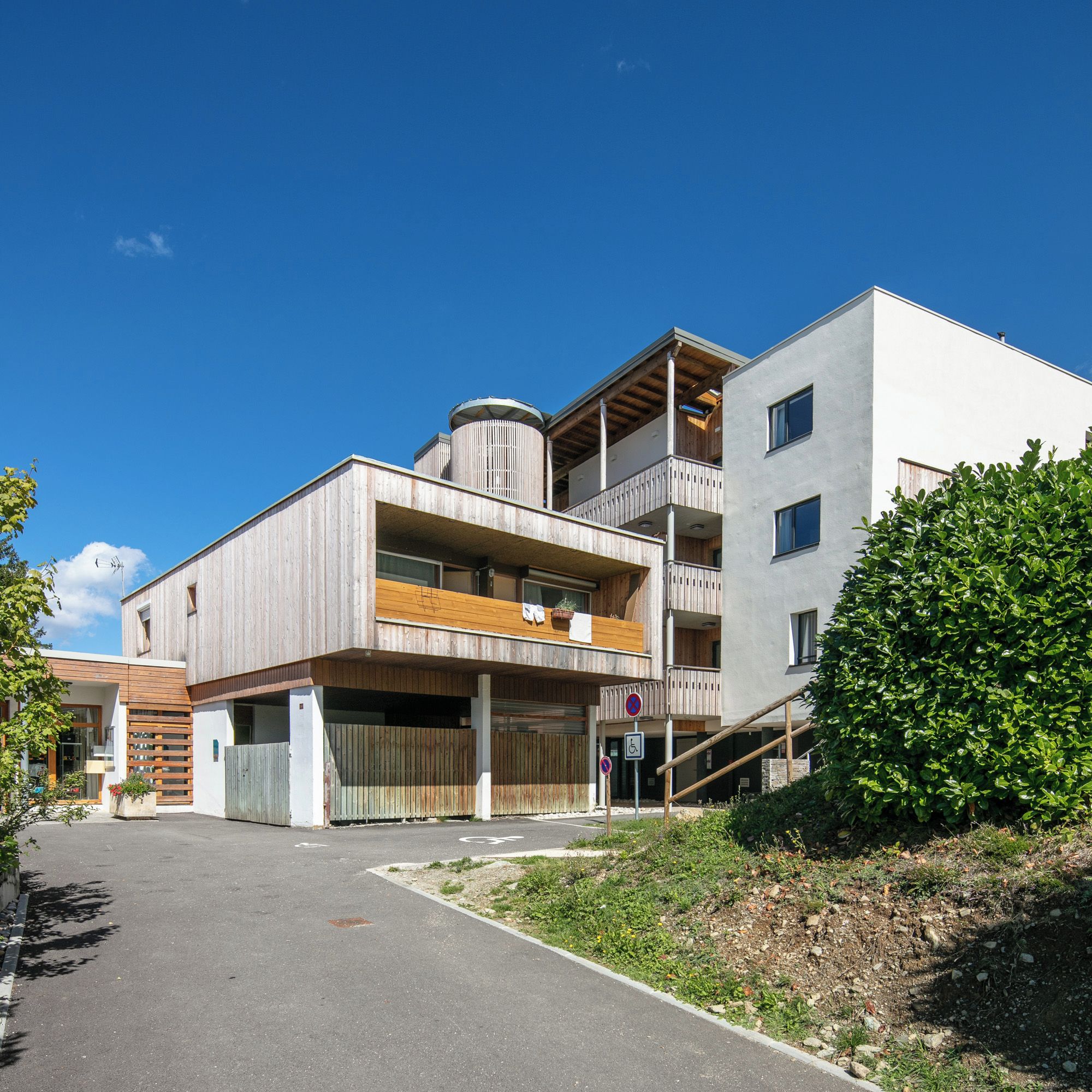 Un edificio moderno de varios pisos con techo plano, un tanque de agua cilíndrico y un área exterior cubierta con bancos de madera.