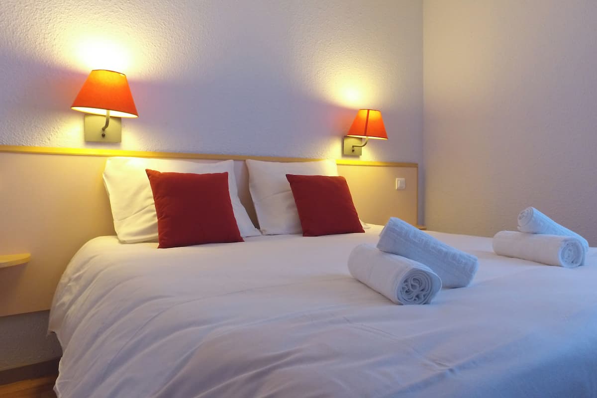 A hotel room with a bed, two red pillows, two white pillows, two wall-mounted lamps, and rolled towels on the bed.