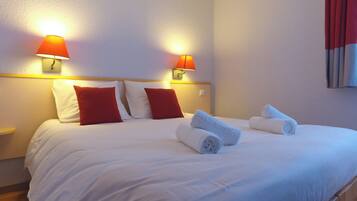 A hotel room with a bed, two red pillows, two white pillows, two wall-mounted lamps, and rolled towels on the bed.