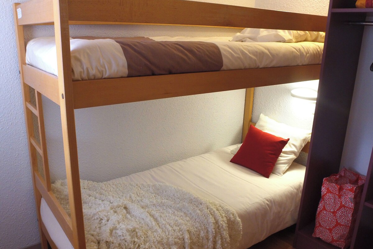 Ein Stockbett mit einer unteren Liege, einer weißen Decke, einem roten Kopfkissen und einer Holzleiter.