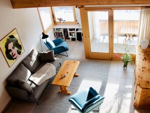 Ein Wohnzimmer mit einer Couch, einem Couchtisch und einem blauen Sessel. Ein Bücherregal und eine Tür, die zu einem Außenbereich führen.