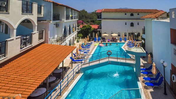 A pool area with blue lounge chairs, a diving board, and a staircase leading to a building with a red-tiled roof. - Vossos (Zakynthos)
