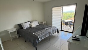 Une chambre avec un lit, un ventilateur de plafond, un bureau avec une télévision et une porte-fenêtre coulissante donnant sur un balcon.