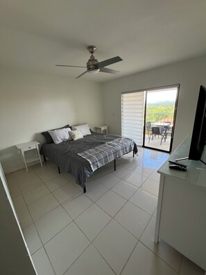 A bedroom with a bed, a ceiling fan, a desk with a TV, and a sliding glass door to a balcony. - Terrific 2-bedroom apartment with AC in Palmas Del Mar with Breathtaking views (Palmas del Mar)