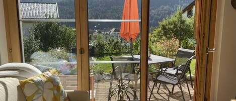 A room with a sliding glass door leading to an outdoor patio with a table and chairs, a sofa with a cushion, and a potted plant.