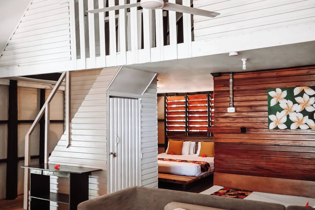 Ein Schlafzimmer im Loft-Stil mit schrägem Dach, einem Bett mit orangefarbener Bettwäsche, einem Holz-Kopfende und einem Deckenventilator.