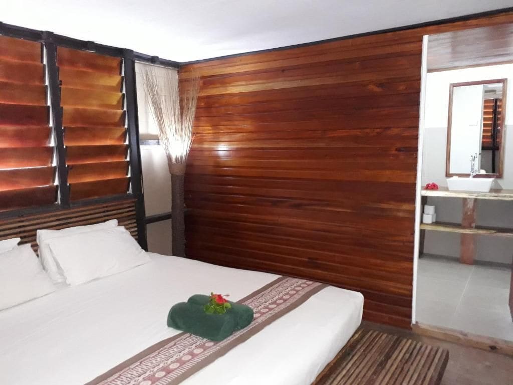 Ein Hotelzimmer mit Holzwand, einem Bett mit weißen Leinentüchern, einem grünen Zierkissen und einem Badezimmer mit Spiegel und Waschbecken.