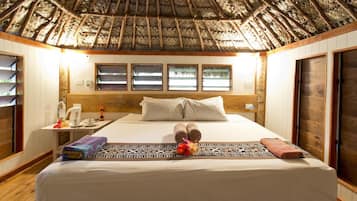 Habitación de hotel con techo de paja, una cama con almohadas, una mesita de noche con una taza y una ventana con vistas.