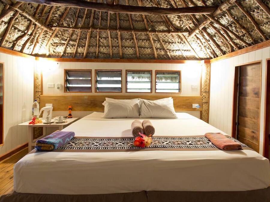 A hotel room with a thatched roof, a bed with pillows, a bedside table with a cup, and a window with a view.
