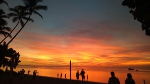 A beach scene with people silhouetted against a sunset, palm trees, and a calm sea. - Likuri Island Resort Fiji (Lomowai)
