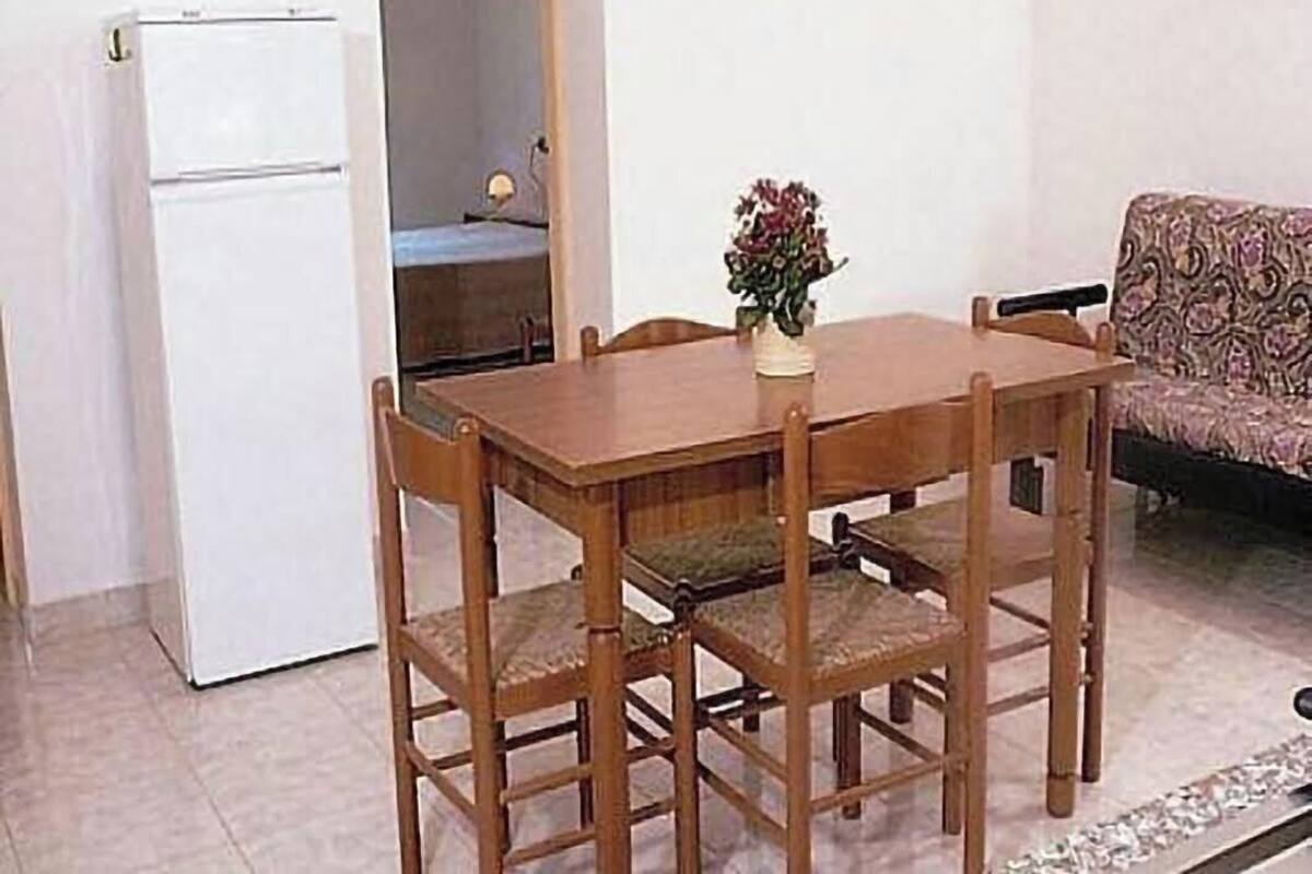 A dining table with four chairs, a small vase with flowers, a white refrigerator, and a patterned sofa in a room with tiled flooring.