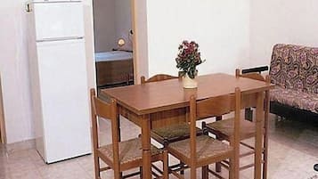 Un tavolo da pranzo con quattro sedie, un piccolo vaso con fiori, un frigorifero bianco e un divano con motivo in una stanza con pavimento piastrellato.