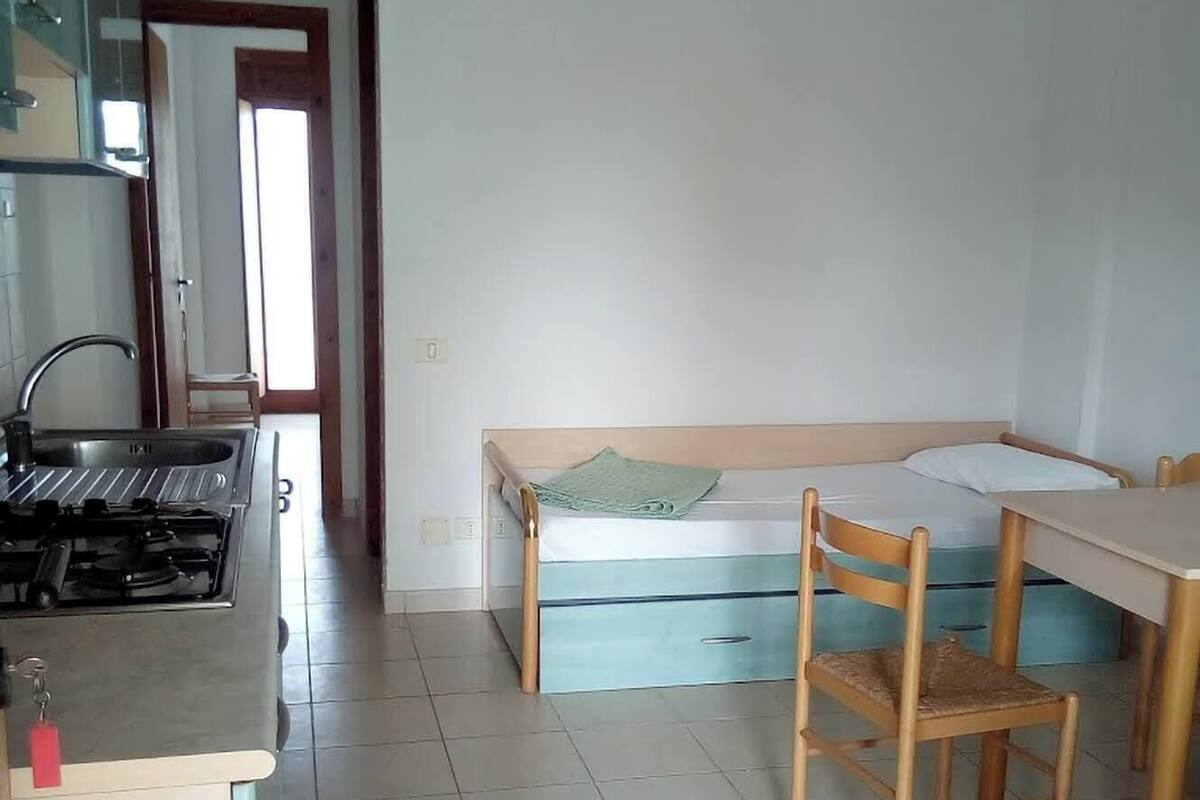 A small, clean room with a kitchenette, a bed with a green pillow, and a wooden chair.