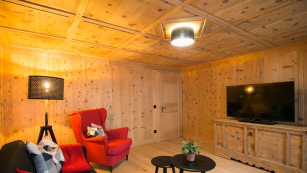 Ein Zimmer mit Holzwänden, einer Couch, zwei roten Sesseln, einem Fernseher und einer Lampe.