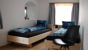 Ein Schlafzimmer mit zwei Einzelbetten, einem Holztisch, einem Stuhl, einem kleinen Fenster mit Vorhängen und einer Wandleuchte.
