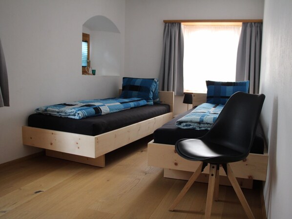 A bedroom with two single beds, a wooden desk, a chair, a small window with curtains, and a wall-mounted light fixture.