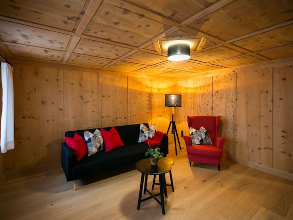 A room with wooden walls and ceiling, a black sofa with red and patterned cushions, a red armchair, a small round table with a plant, and a tripod lamp.