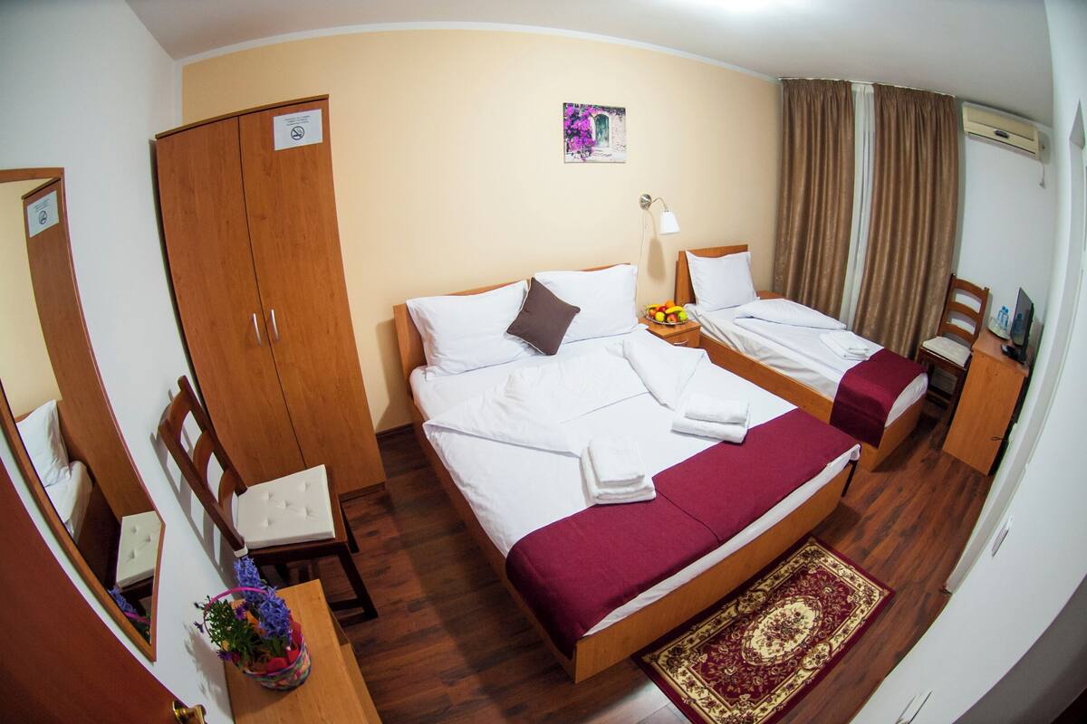 A hotel room with a large bed, a wooden wardrobe, a desk with a chair, a small table with a vase of flowers, and a wall-mounted air conditioner.