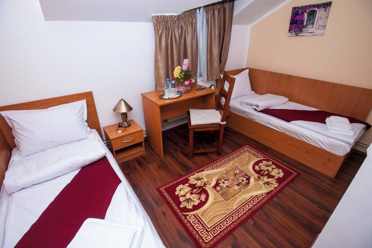 A hotel room with two single beds, a wooden desk, a chair, a small table, a lamp, and a decorative rug.