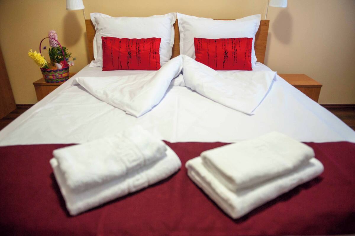 A neatly made bed with white linens and red pillows, a basket of flowers, and folded towels on a red bedspread.