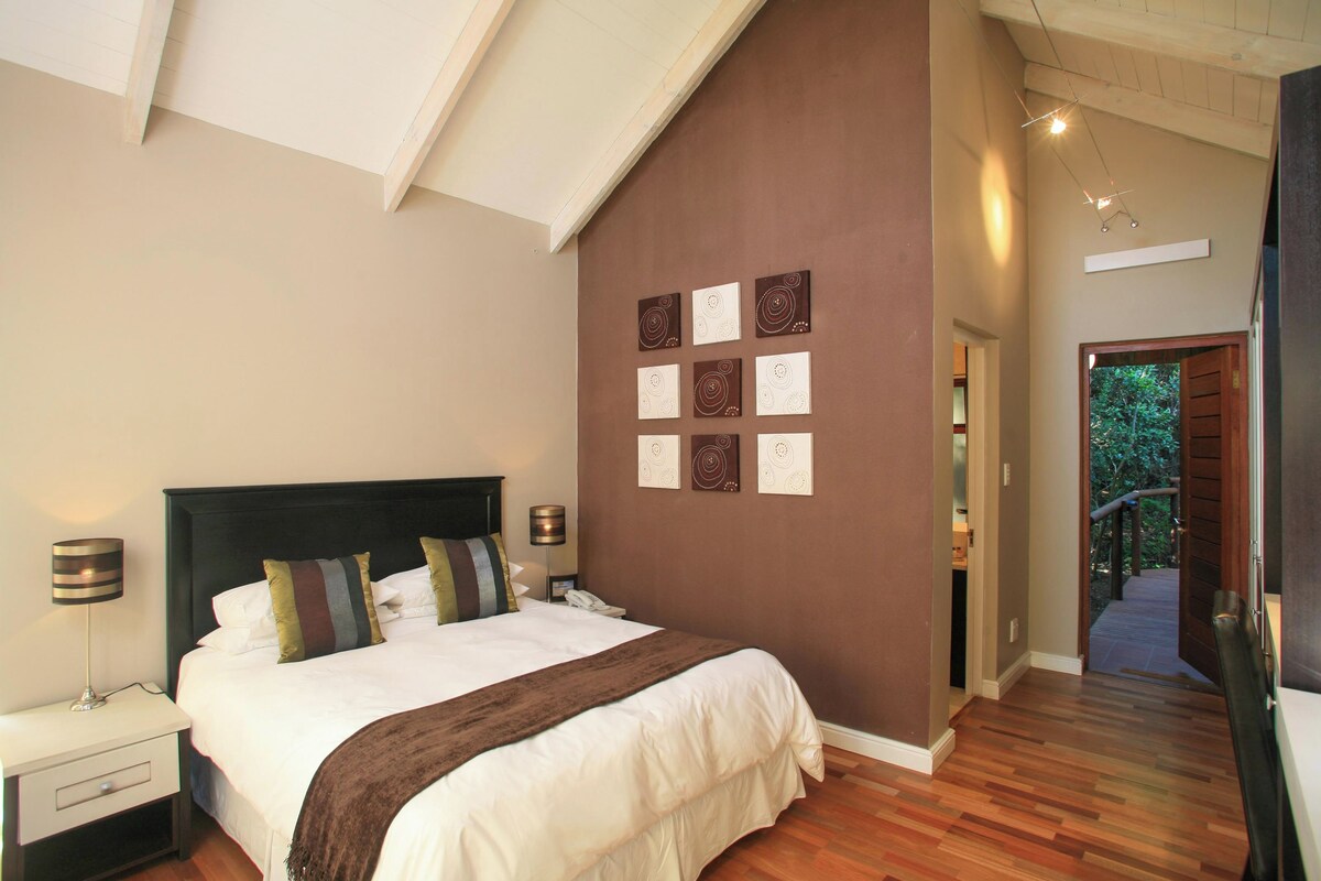 A bedroom with a large bed, a nightstand, a lamp, and a wall with framed artwork.