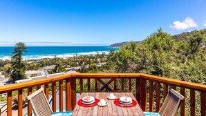 A wooden deck with a table set for two, overlooking a coastal landscape with a view of the ocean and hills. - Under the Boardwalk Guest House (Wilderness)