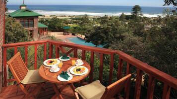 Terraza de madera con una mesa dispuesta para el desayuno, con vista a la playa y al océano.