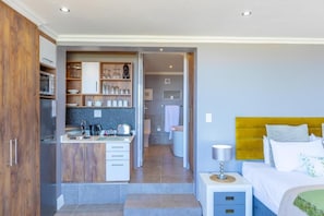 A compact hotel room with a kitchenette, a bed with a yellow headboard, and a small bedside table.