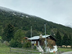 Eine Holzhütte mit Metalldach, umgeben von einem Holzzaun, vor einer verschneiten Berglandschaft und dichtem Wald.