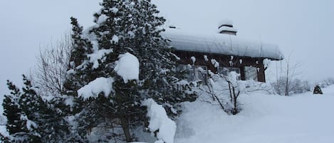 Un edificio cubierto de nieve con techo inclinado, rodeado de árboles y arbustos cargados de nieve.