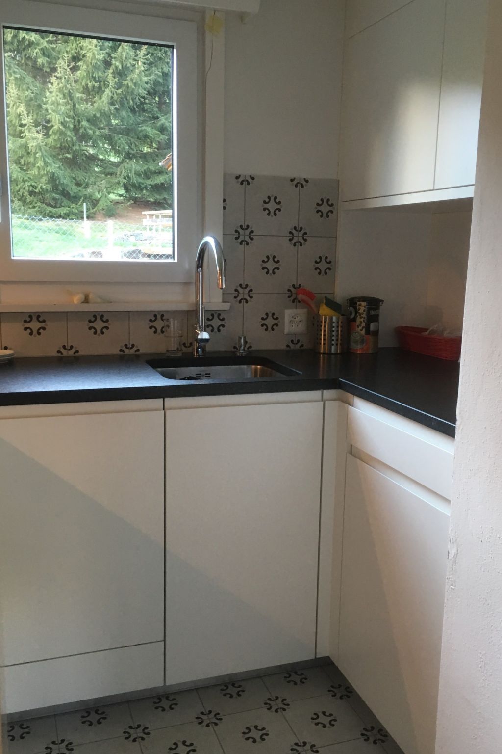 A kitchen with white cabinets, a black countertop, a window with a view of greenery, and a tiled backsplash.