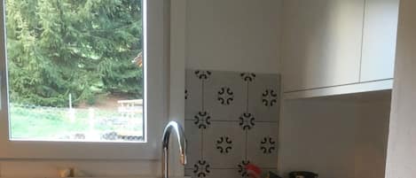 Una cocina con armarios blancos, encimera negra, ventana con vistas a zonas verdes y salpicadero de azulejos.