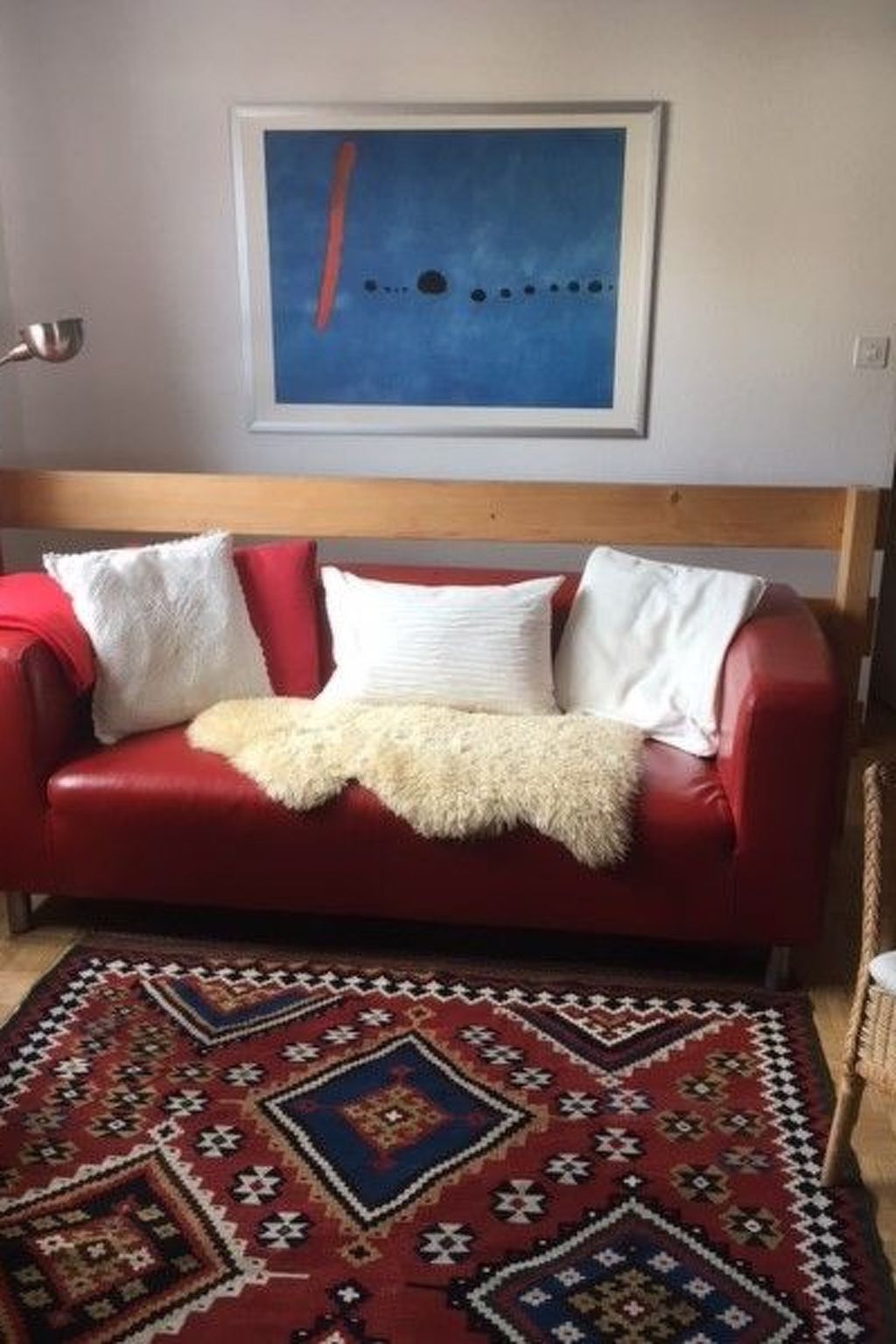 Un sofá de cuero rojo con cojines blancos y una manta beige, un tapete estampado y un cuadro abstracto azul en la pared.
