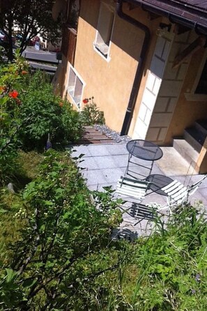 A garden area with a metal chair and a table, surrounded by greenery and a building with a window and door. - Ferienhaus Chasa Silva, (Lüsai) by Interhome (Lü)