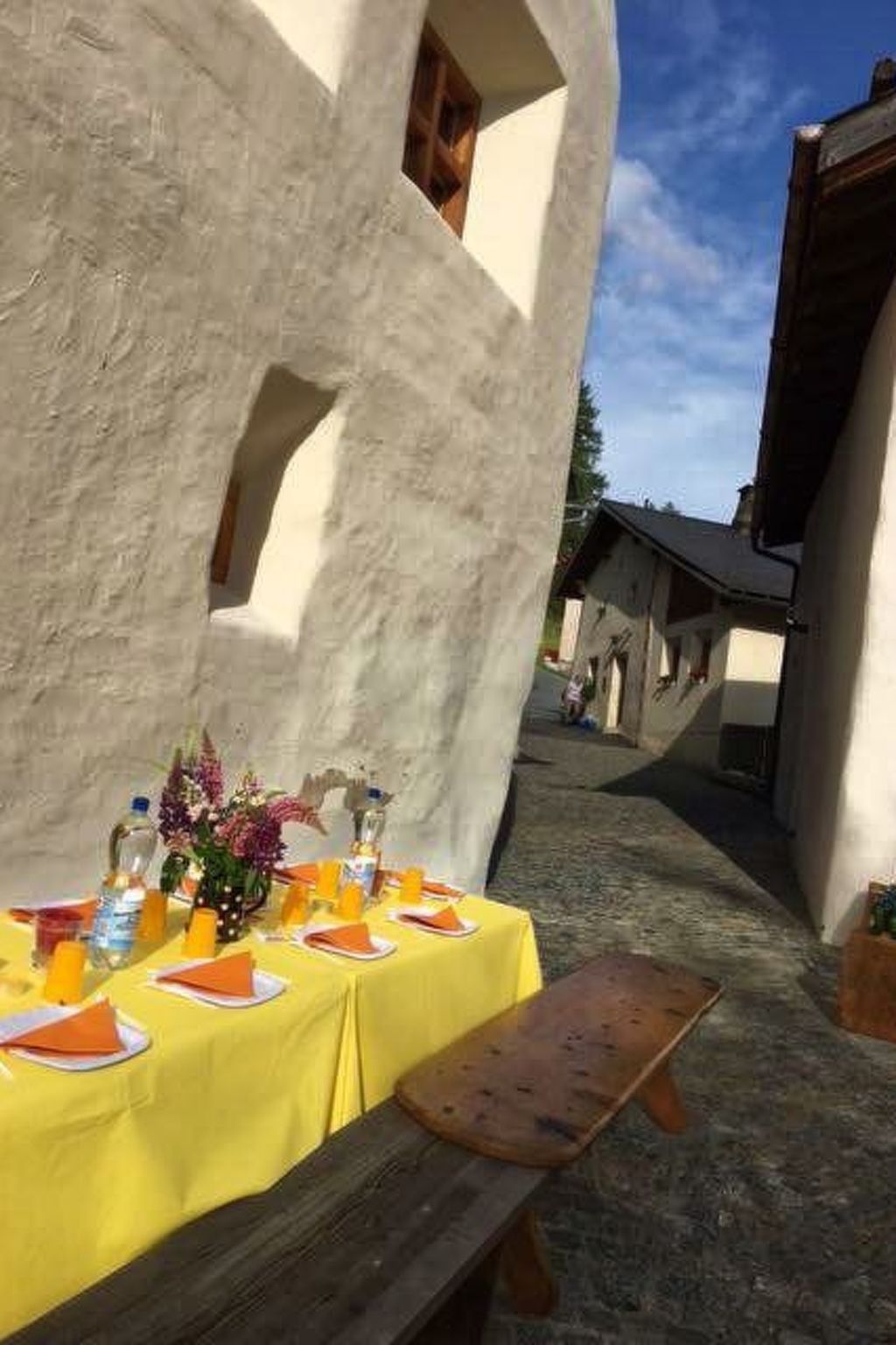 Taula de menjador a l'aire lliure amb estovàtel daurat, una panxa de fusta i vista de construccions tradicionals al fons.
