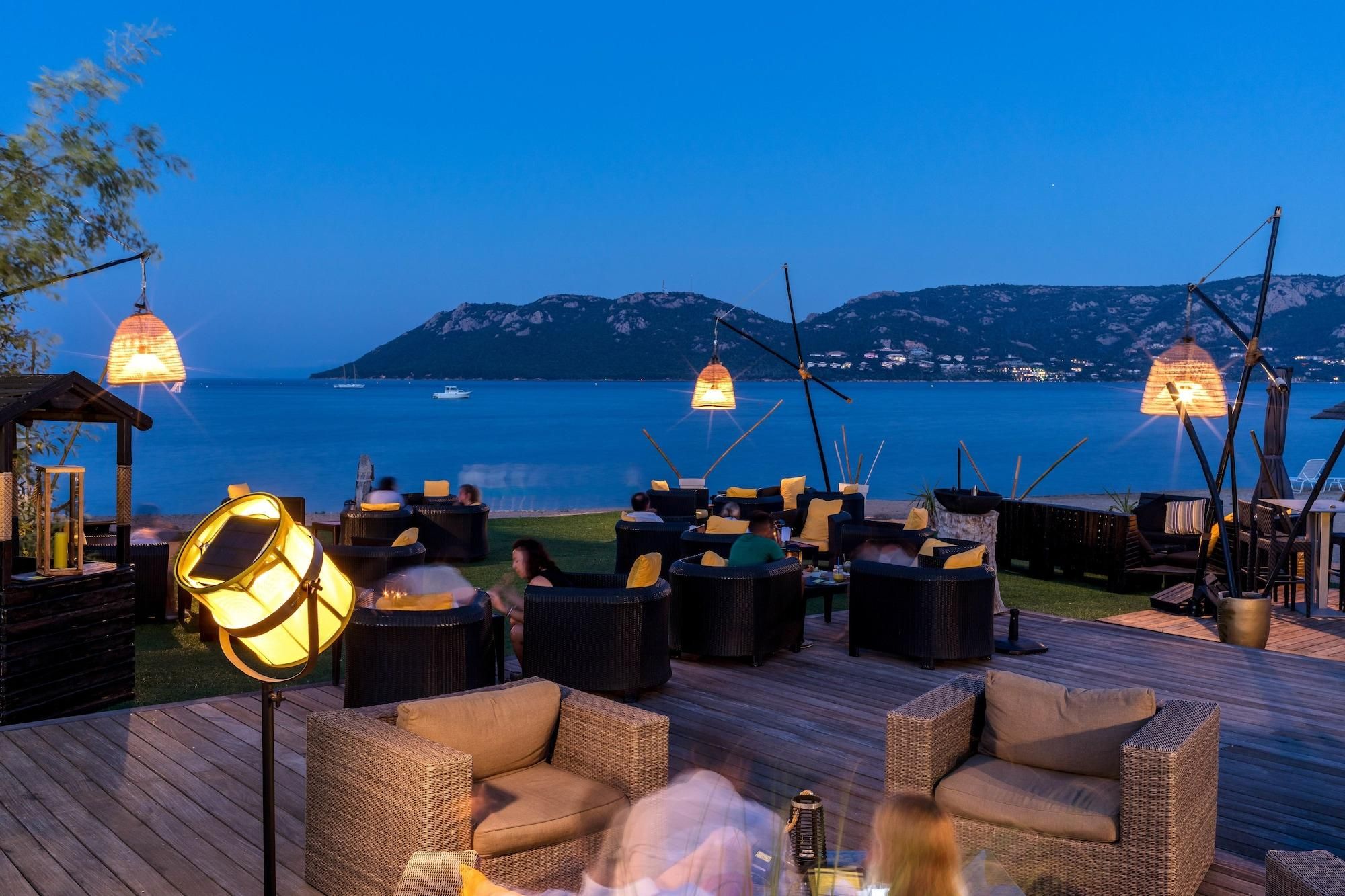An outdoor seating area with wicker furniture and hanging lamps, overlooking a body of water and distant mountains.