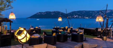 An outdoor seating area with wicker furniture and hanging lamps, overlooking a body of water and distant mountains.