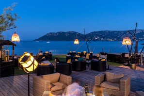 An outdoor seating area with wicker furniture and hanging lamps, overlooking a body of water and distant mountains. - Paese Serenu (Porto-Vecchio)