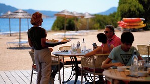 Un restaurante junto a la playa con terraza, un camarero sirviendo un plato, y vista al mar y las montañas al fondo.