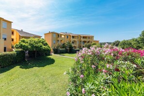 A courtyard with a well-maintained lawn, a flower bed with pink flowers, and a yellow building with multiple balconies. - Paese Serenu (Porto-Vecchio)