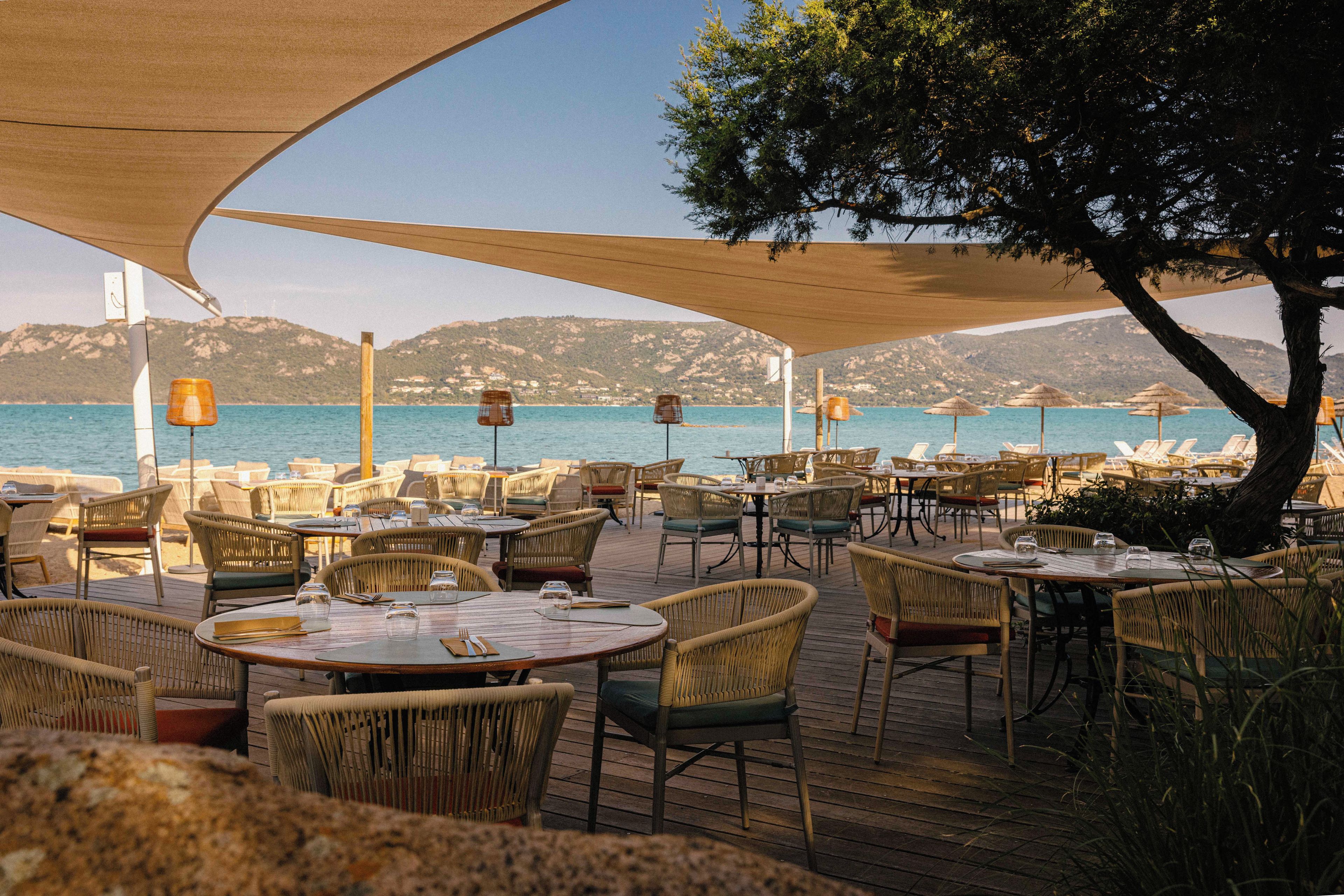 An outdoor dining area with wicker furniture and tables, overlooking a body of water and hills.