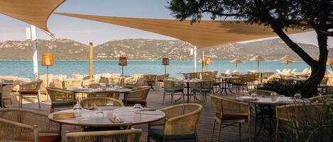 An outdoor dining area with wicker furniture and tables, overlooking a body of water and hills.