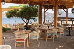 An outdoor dining area with wooden tables and wicker chairs, overlooking a body of water and mountains. - Paese Serenu (Porto-Vecchio)