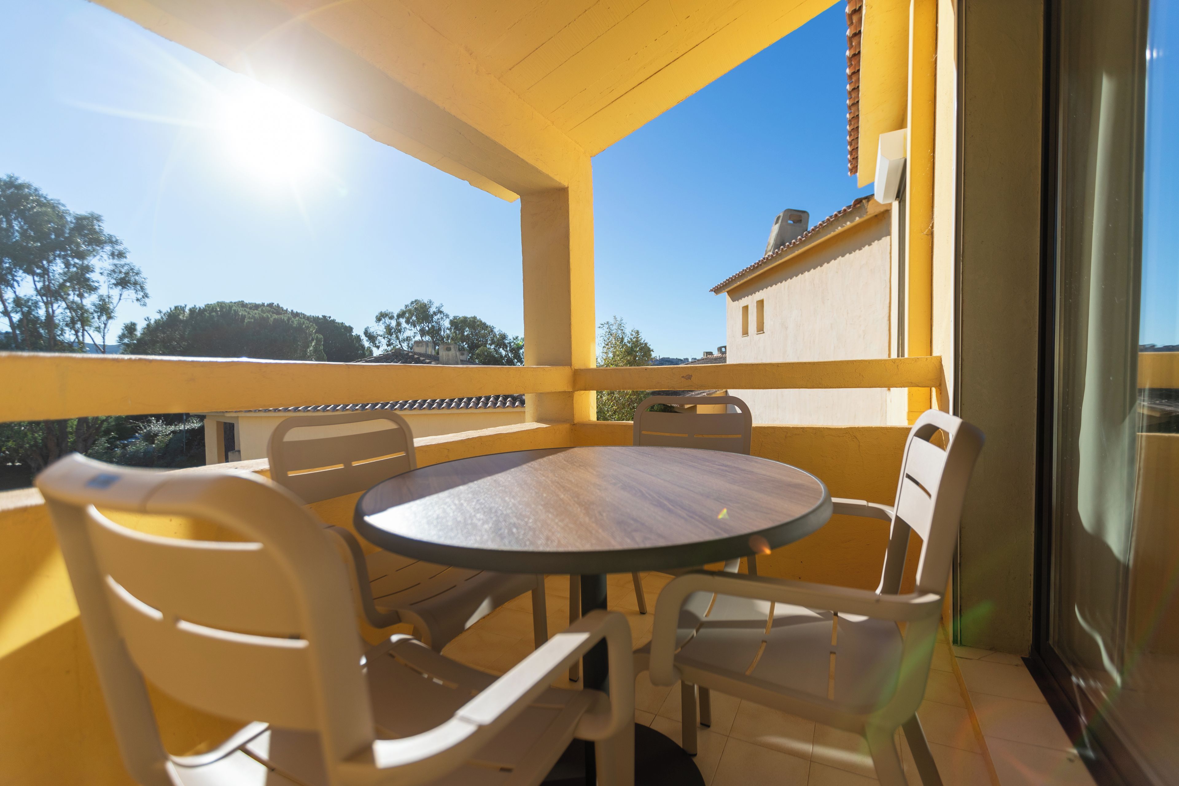 A balcony with a round table and chairs, a view of trees and buildings, and a clear sky.