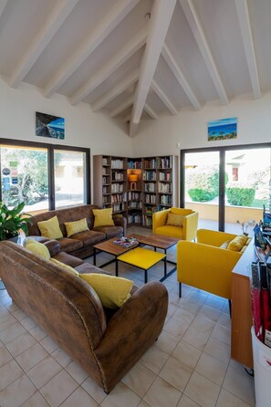 Uma sala de estar ampla, com um sofá de couro marrom, poltronas amarelas, uma mesa de centro de madeira e uma estante repleta de livros. Janelas grandes oferecem luz natural e vista para o verde lá fora.