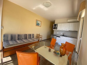 A compact living space with a sofa, dining table, chairs, a kitchen area, and a small dining table with a vase. - Thalassa House Apartments (Agios Vasileios)